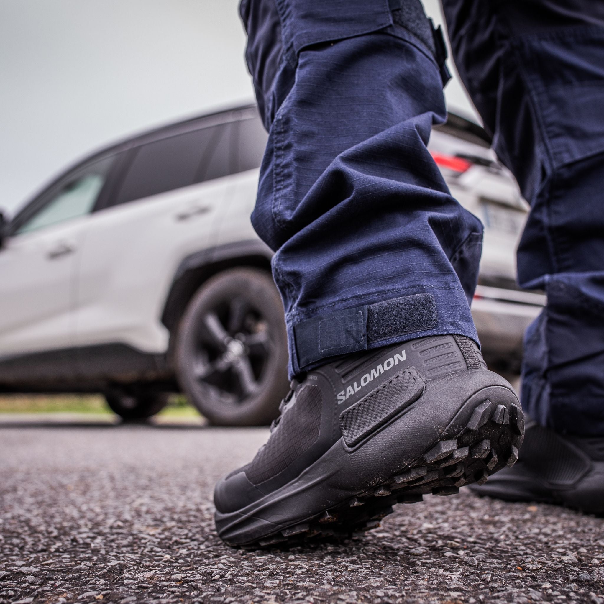 A close-up of a person wearing dark navy pants and black Salomon Genesis boots, standing on asphalt with a white SUV blurred in the background.